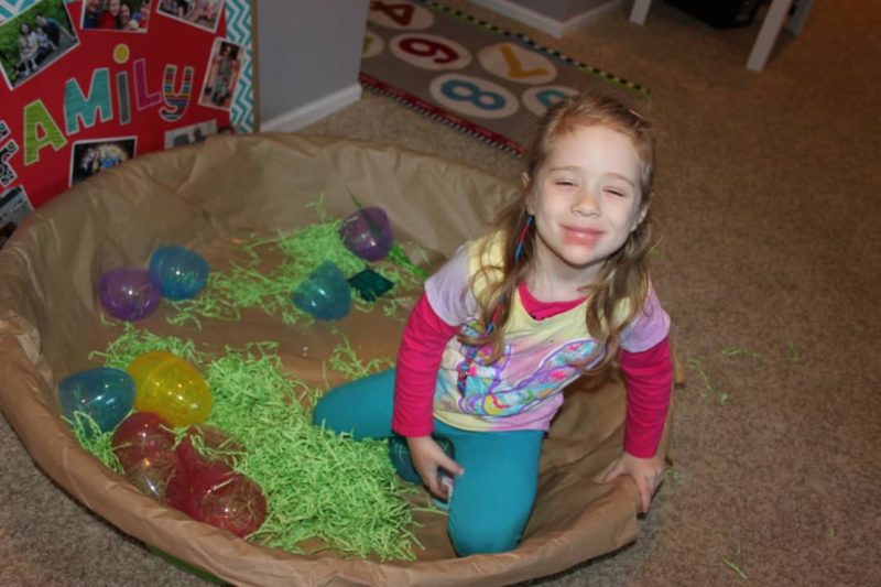 Kiddie pool games can involve role playing like this one that shows a little girl sitting in a kiddie pool covered in brown packing paper. Easter grass and large Easter eggs are inside to make it look like a bird's nest.