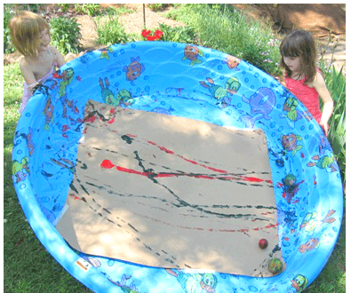 Children stand outside of a kiddie pool with a large piece of cardboard inside of it. Streaks of paint are on the cardboard.
